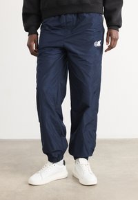 Navyblauwe windbreaker-broek met elastische boorden, voorzien van een witte logo. Gemaakt van lichtgewicht materiaal met een gladde textuur.