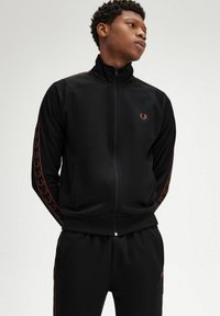 Giacca a track nera con zip e colletto alto, caratterizzata da un discreto logo bordeaux e strisce laterali. Tessuto liscio con tasche sul davanti.