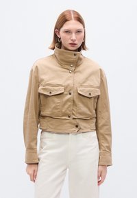 Κοντό μπεζ σακάκι από μαλακό ύφασμα με ψηλό γιακά, δύο μπροστινές τσέπες και κουμπιά με πατιλέτα. Διαθέτει κουτί σχήμα και ραφές λεπτομέρειες.