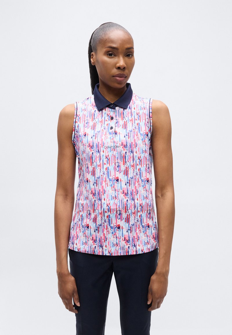 Γυναίκα με μαλλιά δεμένα πίσω φοράει αμάνικη πόλο μπλούζα με φλοράλ σχέδιο και μπλε σκούρο γιακά, καθώς και σκούρο παντελόνι, στέκεται μπροστά σε λευκό φόντο.