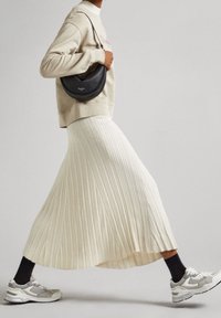 Flödande, plisserad vit kjol i kombination med en beige tröja och grå sportskor. En liten svart läderaxelväska kompletterar outfiten.