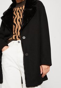 Svart ullblandad kappa med pälskrage, med stora knappar, som bärs över en beige och svart mönstrad tröja och vita byxor.