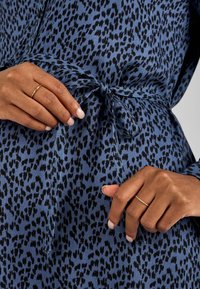 Haljina od plavog materijala s leopard printom s vezanjem u struku. Ima dugačke rukave, uzorak mrlja u crnoj boji i minimalne zlatne prstenaste detalje na prstima.