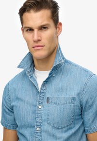 Jasnoniebieska koszula dżinsowa z krótkim rękawem, z kołnierzykiem zapinanym na guziki, kieszenią na piersi i ciemnym przeszyciem. Posiada markową metkę na kieszeni.