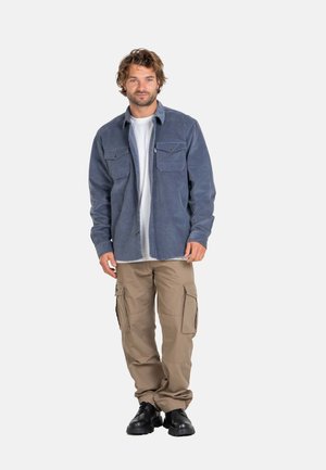 Hombre de pie con una camisa azul de manga larga sobre una camiseta blanca, pantalones cargo beige y zapatos negros sobre un fondo liso.