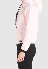 Jasnoróżowa kurtka przeciwwiatrowa z kapturem, czarnymi sznurkami i elastycznymi mankietami. Materiał wydaje się gładki z lekko błyszczącym wykończeniem.