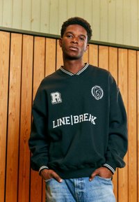 Musta dressipluus, millel on valged kirjateenused "LINE|BREAK" ja logosildid. Omab V-kaelust ja triibulisi varrukaotsasid, kantud helesiniste teksadega.