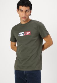 Olivnozelena bombažna majica s kratkimi rokavi, ki ima pravokotni logotip z napisom "Tommy" v beli barvi in "JEANS" v rdeči, okrogla izrezana vratu.