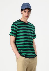 Chemise à rayures vertes et bleu marine à manches courtes avec un col rond et une petite poche poitrine, assortie à un pantalon beige.