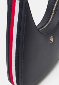 Marineblaue Kunstleder-Schultertasche mit geschwungener Form, ausgestattet mit einem horizontalen Streifen in Rot, Weiß und Blau sowie goldfarbener Logo-Beschläge.