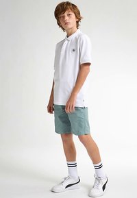 Bílá polokošile s krátkými rukávy a malým logem, doplněná světle tyrkysovými šortkami, bílými ponožkami s černými pruhy a bílými sportovními botami.