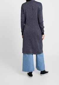 Lång marinblå cardigan med en strukturerad stickdesign. Svarta manschetter och fåll, bärs över vida denimbyxor och svarta sneakers.