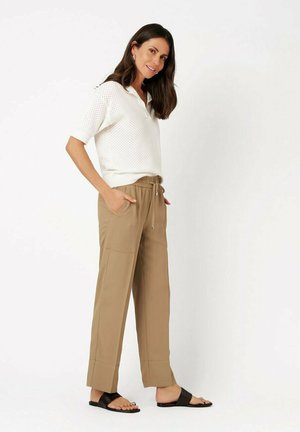 Frau lächelt, trägt ein weißes kurzärmeliges Strick-Polo, beige Hose mit Kordelzug und Taschen sowie schwarze Slip-On-Sandalen, steht vor weißem Hintergrund.