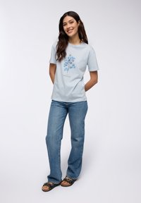 T-shirt di cotone azzurro chiaro con un motivo floreale blu, abbinato a jeans dritti e sandali neri con dettagli decorativi.