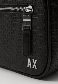 Borsa nera con texture caratterizzata da un logo AX in rilievo, hardware zip in argento e un tirante in pelle. Finitura liscia con dettagli a motivo.