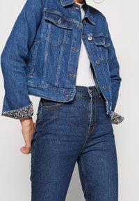 Chaqueta de mezclilla en azul medio con dos bolsillos en el pecho, botones plateados y forro de estampado animal en los puños, combinada con jeans azul a juego.