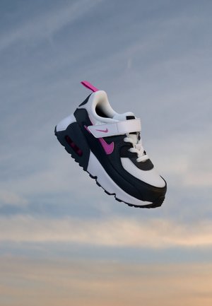Baskets Nike enfant noires, blanches et roses avec fermeture Velcro et lacets flottant devant un ciel nuageux.