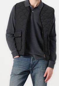 Černá prošívaná vesta se zipem vp�ředu, vybavená dvěma předními kapsami, nošená přes šedé polo tričko s dlouhým rukávem a džínové kalhoty.