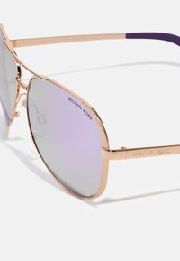 Ochelari de soare aviator cu tonuri de aur și lentile cu gradient violet. Caracteristicile includ un cadru subțire și numele mărcii "Michael Kors" imprimate pe lentile.