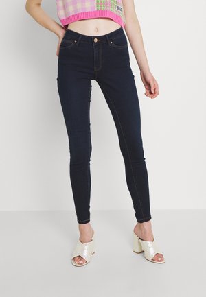 Frau trägt dunkle Skinny-Jeans mit braunen Nähten, einen pink karierten, kurzgeschnittenen Pullover und weiße High-Heel-Sandalen mit Perlendetails.