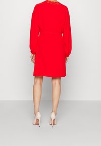 Rotes Kleid mit langen Ärmeln und tailliertem Gürtel. Spitzendetails am Ausschnitt. Getragen mit beige hohen Schuhen.