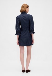 Rochie din denim cu un corsaj mulat, mâneci bufante și manșete cu volane suprapuse. Culoare albastru închis și textură ușoară, stil până la genunchi.