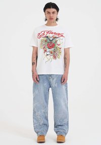 Camiseta gráfica blanca con logo rojo y un diseño colorido estilo tattoo, combinada con unos pantalones vaqueros holgados de patrón azul claro y zuecos marrones.