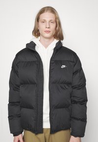 Sort puffer-jakke med lynlås foran, strukturstof og et lille hvidt Nike-logo på øverste venstre side. Båret over en hvid hoodie.
