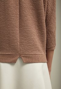 Suéter marrón texturizado con mangas tres cuartos y un dobladillo dividido, combinado con una prenda blanca debajo.
