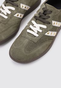 Zapatillas de ante verde oliva con acentos blancos, tachuelas doradas y cordones marrones en contraste. Con una suela de goma texturizada.