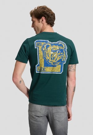 Homme aux cheveux bruns et tatouages portant un t-shirt vert foncé avec un grand logo bulldog usé et le texte "LEE 89" au dos.