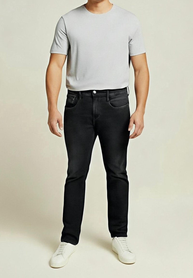 Mężczyzna ubrany w jasnoszarą koszulkę z krótkim rękawem, czarne dopasowane dżinsy i białe trampki, stojący na tle jednolitego beżowego tła.