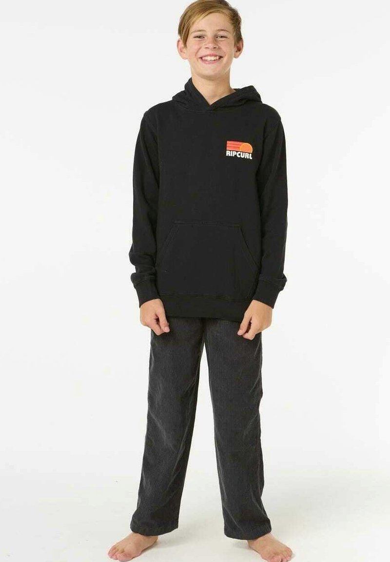Haut à capuche noir avec une poche kangourou à l'avant, arborant un logo "Ripcurl" coloré. Associé à un pantalon sombre et texturé. Modèle debout pieds nus.