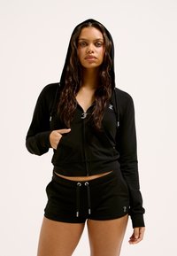 Schwarze Zip-Hoodie mit Kapuze und Fronttasche, kombiniert mit passenden schwarzen Shorts mit Kordelzug und Logodetails. Glatte Stoffoberfläche.