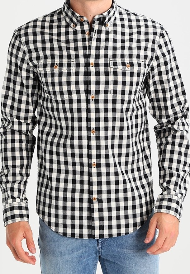 Homme portant une chemise à carreaux noir et blanc à manches longues avec des boutons marron et un jean bleu clair, debout sur un fond blanc.