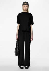 T-shirt nera cropped e pantaloni a gamba larga in materiale morbido, abbinati a scarpe nere e un cappellino nero, mentre tiene una borsa nera.