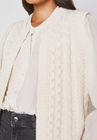 Gilet en tricot crème avec des motifs en relief, des bords côtelés et un col rond, superposé à une blouse légère et un jean foncé.