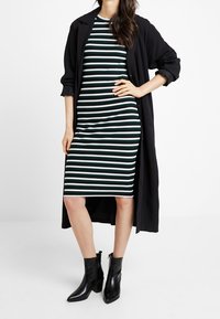 Schwarzer langer Mantel über einem grün-weißen gestreiften, gerippten Kleid, knöchellang. Schwarze Blockabsatzstiefel mit strukturierter Oberfl che.