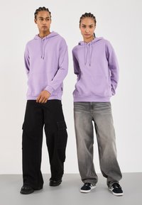 Due persone indossano felpe leggere viola coordinate con tasche a marsupio. Un paio indossa pantaloni cargo neri, l'altro jeans grigi chiari e scarpe da ginnastica nere.
