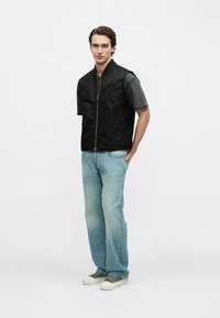 Schwarze ärmellose Steppweste mit Reißverschluss und Logodetail, kombiniert mit hellblauen Jeans und olivgrünen Turnschuhen. Glatte Textur.