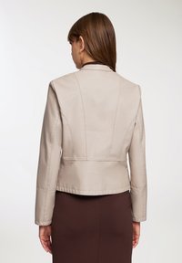 Giacca in pelle beige con un design strutturato, caratterizzata da un colletto alto, maniche lunghe e dettagli cuciti. Indossata sopra una gonna marrone.