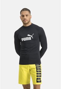 Schwarzes Langarm-Rashguard mit weißem "PUMA"-Logo, kombiniert mit knielangen, hellgelben Shorts, die an der Seite schwarzen "PUMA"-Text haben.