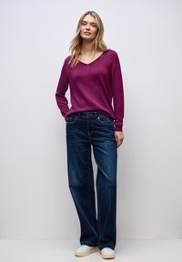 Pull-over long de couleur violette avec un décolleté en V, associé à un jean large bleu foncé et des baskets blanches. Texture lisse, design décontracté et coupe relaxée.