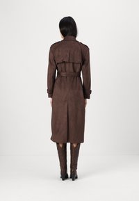 Brązowy zamszowy trencz z paskiem w talii, naramiennikami i warstwowym tyłem; noszony z brązowymi, teksturowanymi botkami do kolan.