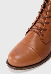 Bottine en cuir marron avec un bout arrondi, surface lisse et détails en laçage. Présente un talon en bois empilé à la base.