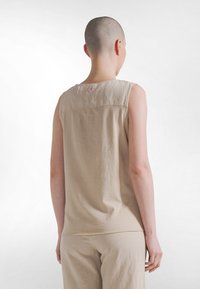 Beige mouwloze top gemaakt van zachte stof, met een naad aan de achterkant en een klein roze labeltje vlakbij de halslijn. Eenvoudig ontwerp met een losse pasvorm.