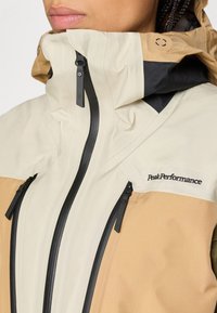 Beige und braune, wetterfeste Jacke mit hohem Kragen, schwarzen Reißverschlüssen und Peak Performance-Logo. Glatte Textur, modernes Design.