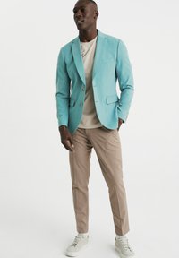Blazer turquesa, camisa beige y pantalones marrones claros. Textura suave, diseño ajustado, botones decorativos y combinado con zapatillas claras.