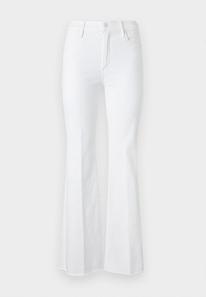 Noor naine, kes kannab tumesinist varrukateta topi, valgeid laiade säärega teksapükse, mille ääred on katki hõõrutud, ning musti kõrgete kontsadega sandaale, seisab hele taustaga ees.