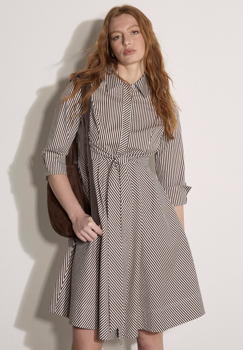 Femme aux longs cheveux roux portant une robe rayée verticale marron et blanche avec une ceinture nouée, tenant un sac à épaule marron, debout contre un mur blanc.